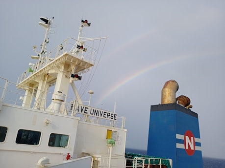 A ship's structure with visible antennae and equipment is set against a cloudy sky with a faint rainbow arcing above. Part of the ship is labeled 'NAVE UNIVERSE' and features a large funnel displaying a red circle with an 'N' on it.