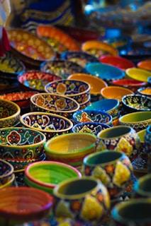 A vibrant display of intricately patterned ceramic bowls in various colors, arranged in layers. The ceramics feature detailed floral and geometric designs, each bowl showcasing a unique pattern and color scheme.