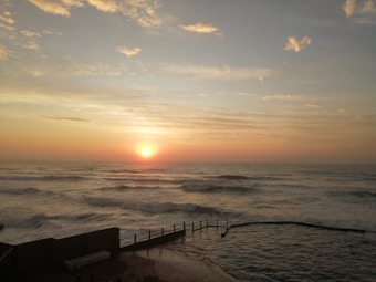 A serene sunrise over the ocean, with soft waves and a clear horizon. The sun casts warm orange and pink hues across the sky, blending into light blue and soft yellow tones. A swimming pool is visible on the right, bordered by a fence, with the sea and sky stretching beyond.