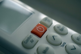 A close-up view of a remote control, highlighting a red power button surrounded by several white buttons with various functions such as temperature control and fan speed. The display screen is mostly off, reflecting some light.