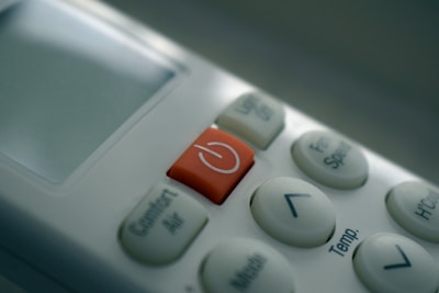 A close-up view of a remote control, highlighting a red power button surrounded by several white buttons with various functions such as temperature control and fan speed. The display screen is mostly off, reflecting some light.