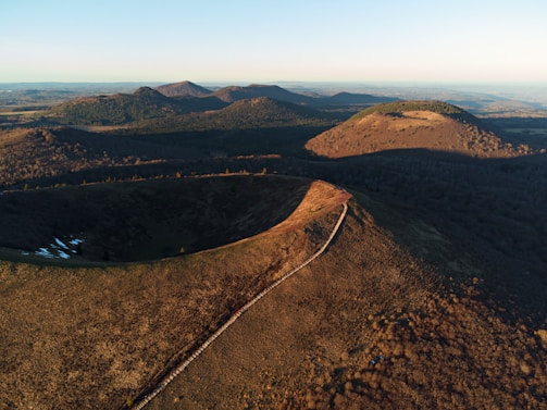 An aerial view of a volcanic landscape features a series of rounded hills and a distinct crater formation in the foreground. The terrain is covered with sparse vegetation, highlighting the earthy tones of the hills. The area is illuminated by the warm light of either sunrise or sunset, casting long shadows across the slopes.