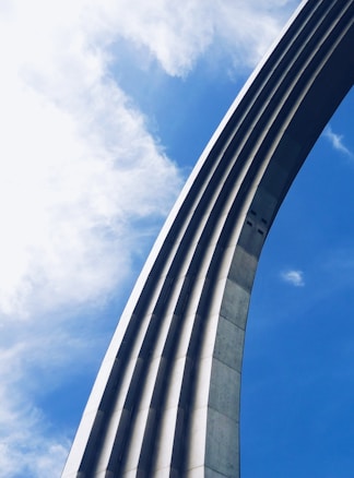 A metallic arch takes up a significant portion of the right side, with its sleek lines curving against the clear blue sky. Fluffy white clouds gently blend with the sky, creating a contrast to the industrial structure.