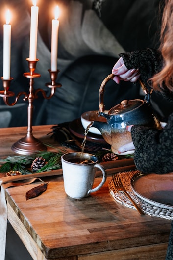 A cozy scene with a person pouring tea from a teapot into a mug on a wooden table. The table is adorned with a plate, pine cones, a leaf, and green foliage. A candelabra with lit candles adds warmth to the atmosphere.