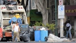 Several individuals are engaged in loading trash into a garbage truck on a city street. The scene includes large black garbage bags, blue barrels, and a no parking sign. Nearby, a man in a white tank top holds a beverage, while another in a yellow shirt stands on the truck. A person walks by with a backpack, near a building and some trees.