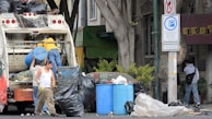 Several individuals are engaged in loading trash into a garbage truck on a city street. The scene includes large black garbage bags, blue barrels, and a no parking sign. Nearby, a man in a white tank top holds a beverage, while another in a yellow shirt stands on the truck. A person walks by with a backpack, near a building and some trees.