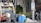 Several individuals are engaged in loading trash into a garbage truck on a city street. The scene includes large black garbage bags, blue barrels, and a no parking sign. Nearby, a man in a white tank top holds a beverage, while another in a yellow shirt stands on the truck. A person walks by with a backpack, near a building and some trees.