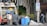 Several individuals are engaged in loading trash into a garbage truck on a city street. The scene includes large black garbage bags, blue barrels, and a no parking sign. Nearby, a man in a white tank top holds a beverage, while another in a yellow shirt stands on the truck. A person walks by with a backpack, near a building and some trees.