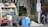 Several individuals are engaged in loading trash into a garbage truck on a city street. The scene includes large black garbage bags, blue barrels, and a no parking sign. Nearby, a man in a white tank top holds a beverage, while another in a yellow shirt stands on the truck. A person walks by with a backpack, near a building and some trees.