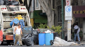 Several individuals are engaged in loading trash into a garbage truck on a city street. The scene includes large black garbage bags, blue barrels, and a no parking sign. Nearby, a man in a white tank top holds a beverage, while another in a yellow shirt stands on the truck. A person walks by with a backpack, near a building and some trees.