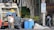 Several individuals are engaged in loading trash into a garbage truck on a city street. The scene includes large black garbage bags, blue barrels, and a no parking sign. Nearby, a man in a white tank top holds a beverage, while another in a yellow shirt stands on the truck. A person walks by with a backpack, near a building and some trees.