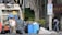Several individuals are engaged in loading trash into a garbage truck on a city street. The scene includes large black garbage bags, blue barrels, and a no parking sign. Nearby, a man in a white tank top holds a beverage, while another in a yellow shirt stands on the truck. A person walks by with a backpack, near a building and some trees.