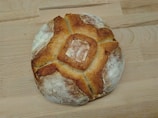 A round, rustic loaf of bread with a crusty, golden-brown exterior and a dusting of flour on top. The pattern on the bread suggests a traditional baking method, and it is resting on a light wooden surface.