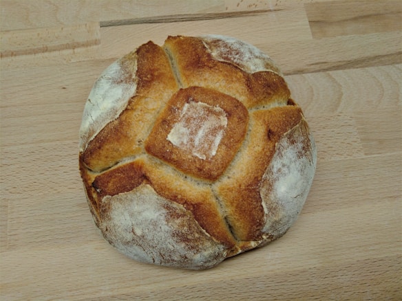 A round, rustic loaf of bread with a crusty, golden-brown exterior and a dusting of flour on top. The pattern on the bread suggests a traditional baking method, and it is resting on a light wooden surface.
