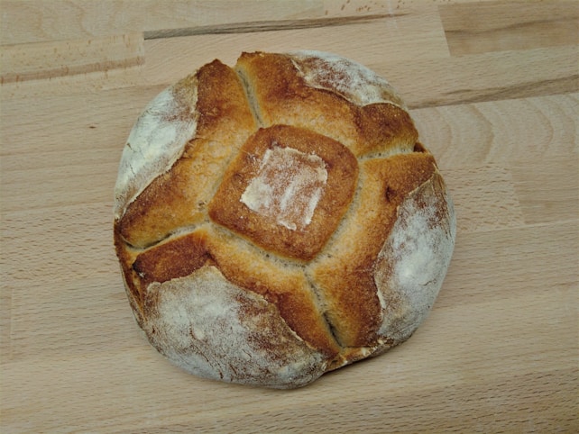 A round, rustic loaf of bread with a crusty, golden-brown exterior and a dusting of flour on top. The pattern on the bread suggests a traditional baking method, and it is resting on a light wooden surface.