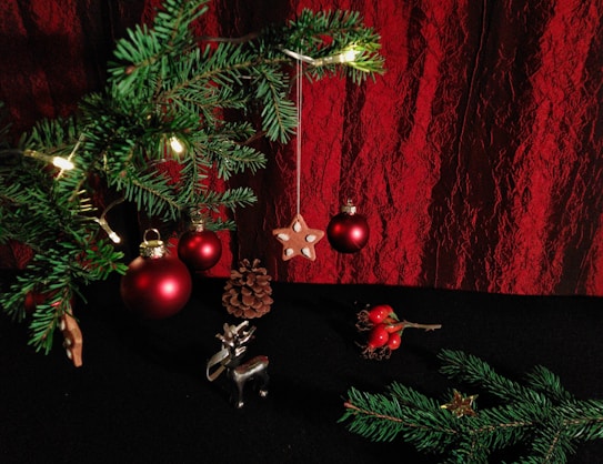 A festive setting featuring a branch of a Christmas tree adorned with red baubles, small lights, and a hanging star ornament. A pine cone, a silver reindeer figure, and red berries on a small branch are placed on a dark surface. The background consists of red textured drapery.