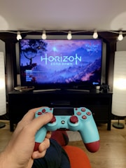 A person is holding a blue and maroon PlayStation controller, facing a television displaying the game Horizon Zero Dawn. The TV is set on a black stand, with decorative string lights above it and lamps on either side. The room has a cozy ambiance with a wooden floor.