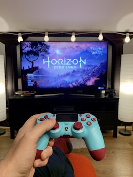 A person is holding a blue and maroon PlayStation controller, facing a television displaying the game Horizon Zero Dawn. The TV is set on a black stand, with decorative string lights above it and lamps on either side. The room has a cozy ambiance with a wooden floor.