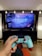 A person is holding a blue and maroon PlayStation controller, facing a television displaying the game Horizon Zero Dawn. The TV is set on a black stand, with decorative string lights above it and lamps on either side. The room has a cozy ambiance with a wooden floor.