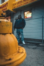 A person stands against a chain-link fence, wearing a dark jacket and jeans. The background features a storefront with a yellow awning adorned with text about food stamps and services. In the foreground, a yellow fire hydrant is prominently visible.