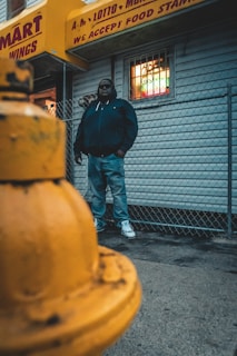 A person stands against a chain-link fence, wearing a dark jacket and jeans. The background features a storefront with a yellow awning adorned with text about food stamps and services. In the foreground, a yellow fire hydrant is prominently visible.