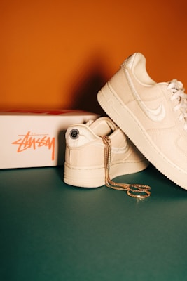 A pair of cream-colored sneakers with a textured finish, featuring a prominent brand logo on the side. They are positioned on a green surface next to a white shoe box branded with orange lettering. A gold chain is casually draped over one of the shoes, adding a touch of elegance.
