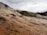 Terraced mineral deposits cascade down a hillside under a cloudy sky, with steam rising in some areas. The formations have a rugged, layered texture and earthy tones, with a backdrop of trees silhouetted against the overcast atmosphere.