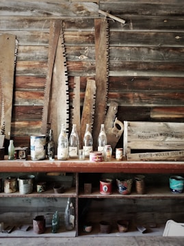 An assortment of vintage saws with rustic wooden handles is arranged against a wooden wall. In front of the saws, there are various glass bottles and old cans displayed on shelves. A wooden crate with text is visible on the right side of the image, contributing to an old-fashioned, industrial ambiance.