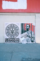 Two posters are displayed on a textured concrete wall. The left poster features a detailed black and white design with a cannabis leaf at the center, accompanied by the text 'Cannabis Justice Now'. The right poster shows a person behind bars and holding a newspaper with a headline that reads 'Marijuana Legalized'. Above them, there's a red signboard with white paint covering previous text.
