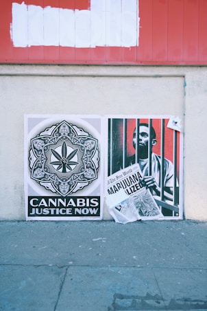 Two posters are displayed on a textured concrete wall. The left poster features a detailed black and white design with a cannabis leaf at the center, accompanied by the text 'Cannabis Justice Now'. The right poster shows a person behind bars and holding a newspaper with a headline that reads 'Marijuana Legalized'. Above them, there's a red signboard with white paint covering previous text.