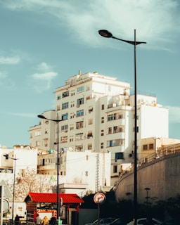 A multi-story residential building with white walls and multiple windows stands under a clear blue sky. In the foreground, there are streetlights, a road sign, and a small structure with a red roof. People can be seen walking near the base of the building.