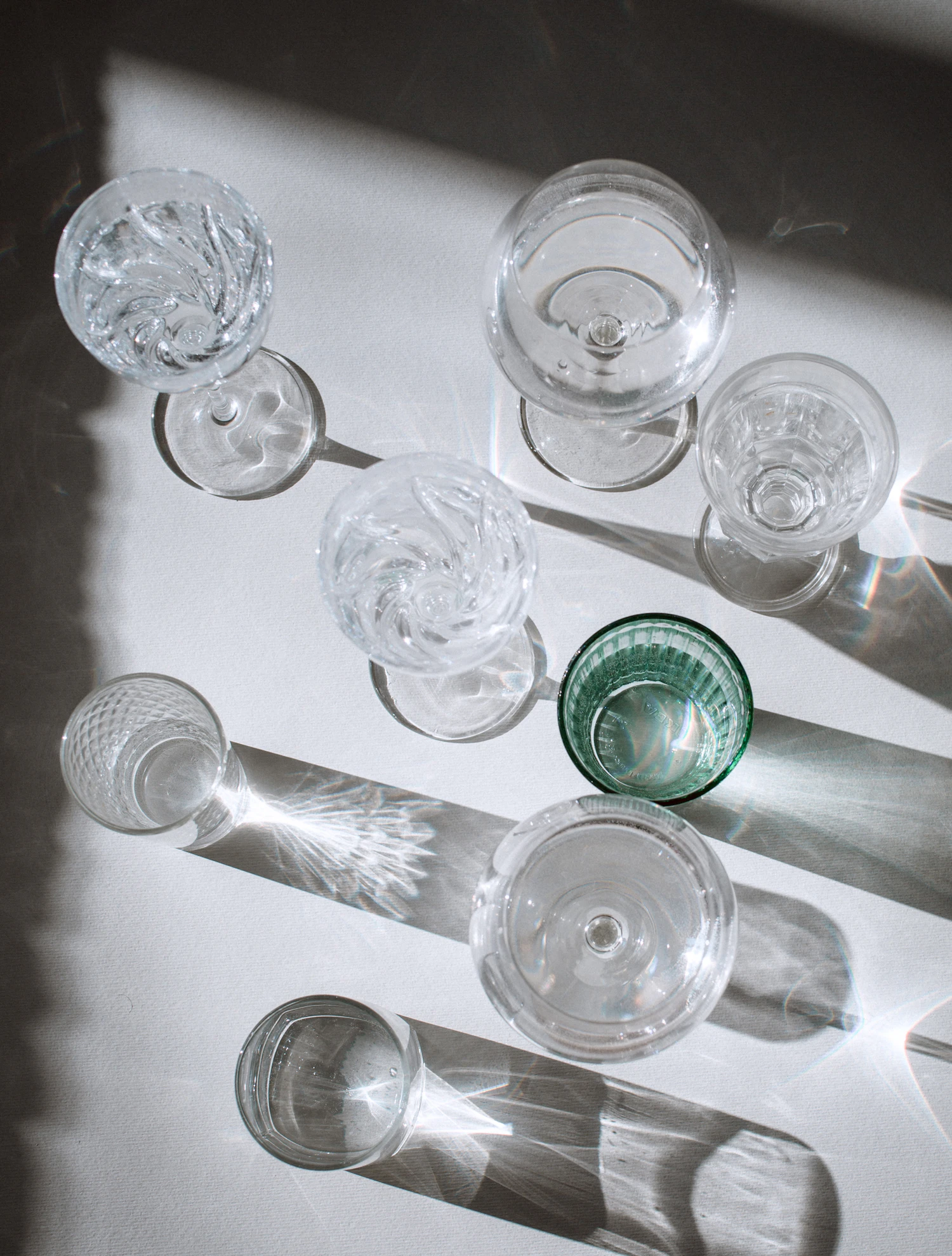 Cut and plain glasses seen from above on a white table, long shadows and a sliver of rainbow at the edge