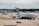 An airport scene features a large passenger aircraft, prominently displaying the Emirates logo, taxiing on the runway. In the background, several other planes from different airlines can be seen parked near the terminal buildings. A ramp worker is directing the aircraft, and various airport equipment is present.