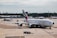 An airport scene features a large passenger aircraft, prominently displaying the Emirates logo, taxiing on the runway. In the background, several other planes from different airlines can be seen parked near the terminal buildings. A ramp worker is directing the aircraft, and various airport equipment is present.