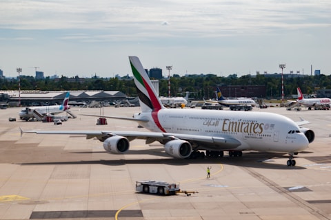 An airport scene features a large passenger aircraft, prominently displaying the Emirates logo, taxiing on the runway. In the background, several other planes from different airlines can be seen parked near the terminal buildings. A ramp worker is directing the aircraft, and various airport equipment is present.