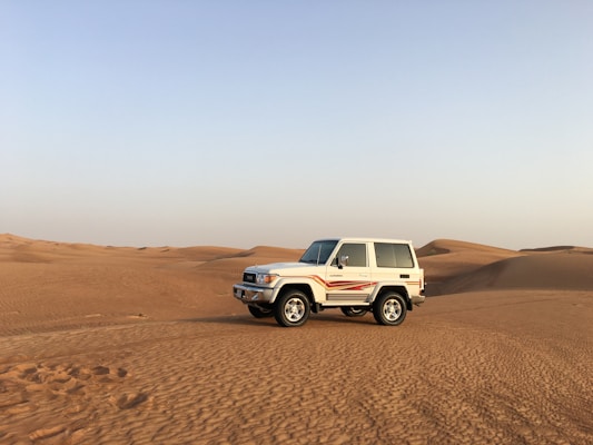 A white SUV with red stripes is parked on expansive, sandy desert dunes under a clear blue sky.