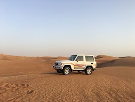 A white SUV with red stripes is parked on expansive, sandy desert dunes under a clear blue sky.