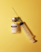 A vial labeled 'COVID-19 Vaccine' and an empty syringe are placed on a yellow background. The vial has a metallic cap, and the syringe appears to be of standard design with measurement markings.