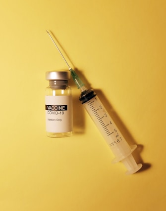 A vial labeled 'COVID-19 Vaccine' and an empty syringe are placed on a yellow background. The vial has a metallic cap, and the syringe appears to be of standard design with measurement markings.