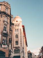 A historic building with a dome and ornate architectural details is prominently featured. The facade includes a vertical sign reading 'Legacy Ottoman'. The sky is clear with a soft, warm light illuminating the structure, enhancing its intricate designs. Adjacent buildings have a more weathered look with noticeable balconies and a banner. The scene captures a blend of historic and urban elements.