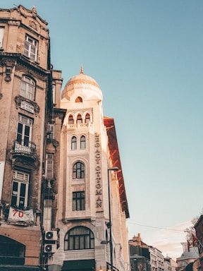 A historic building with a dome and ornate architectural details is prominently featured. The facade includes a vertical sign reading 'Legacy Ottoman'. The sky is clear with a soft, warm light illuminating the structure, enhancing its intricate designs. Adjacent buildings have a more weathered look with noticeable balconies and a banner. The scene captures a blend of historic and urban elements.