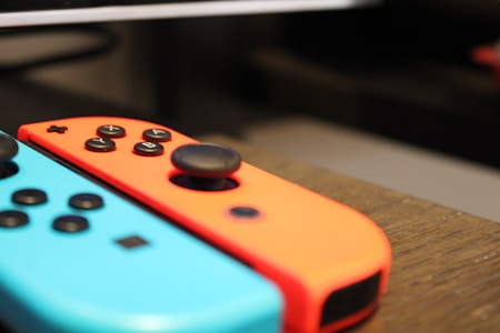 Two handheld gaming controllers with distinct colors, blue and red, resting side by side on a wooden surface. The controllers feature several buttons and a joystick, and are designed with a curved shape for ergonomic holding.