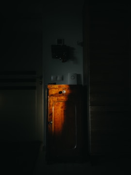 A dimly lit room featuring an old wooden cabinet illuminated by a spotlight. The warm tones of the wood stand out against the dark, shadowy surroundings. Above the cabinet, there are small items and switches mounted on the wall.