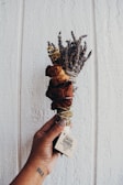 A hand holds a bundle of dried herbs and flowers, including lavender and rose. The bouquet is wrapped with a string and has a small label attached with the text 'Sacred Dwelling.' The background consists of a textured, light-colored wall.