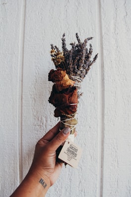 A hand holds a bundle of dried herbs and flowers, including lavender and rose. The bouquet is wrapped with a string and has a small label attached with the text 'Sacred Dwelling.' The background consists of a textured, light-colored wall.