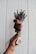 A hand holds a bundle of dried herbs and flowers, including lavender and rose. The bouquet is wrapped with a string and has a small label attached with the text 'Sacred Dwelling.' The background consists of a textured, light-colored wall.