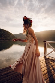 A person in a flowing, elegant dress adorned with floral patterns dances gracefully on a wooden deck by a calm lake, surrounded by mountains. The sun sets in the background, casting a warm glow across the scene. A vibrant flower crown decorates their hair, adding a touch of nature and color to their ensemble.