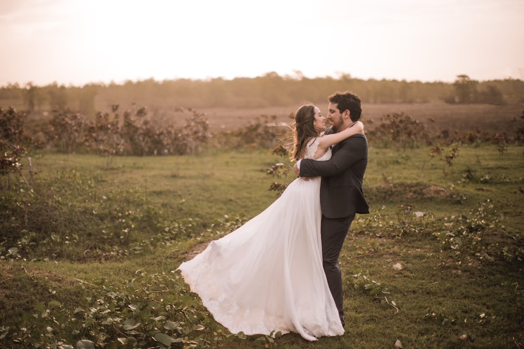woman in white wedding gown kissing man in black suit on green grass field during daytime