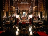 An ornate interior of a traditional temple featuring intricate wooden carvings and golden decorations. The space is filled with various ritual objects, including lanterns, cushions, and altars adorned with offerings. The dim lighting creates a serene and sacred atmosphere.