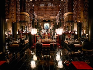 An ornate interior of a traditional temple featuring intricate wooden carvings and golden decorations. The space is filled with various ritual objects, including lanterns, cushions, and altars adorned with offerings. The dim lighting creates a serene and sacred atmosphere.